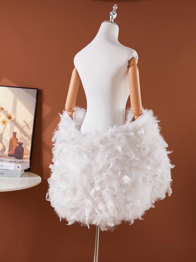 May include: A white dress form with wooden arms is adorned with a skirt made of fluffy white feathers. The dress form is on a silver stand, set against a warm brown background. A framed picture and books are visible in the background.