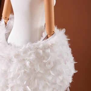 May include: A white, fluffy feather skirt is draped on a mannequin. The skirt is made of soft, white feathers, creating a voluminous and textured appearance. The feathers are densely packed, giving the skirt a full, rounded shape. The background is a solid brown.