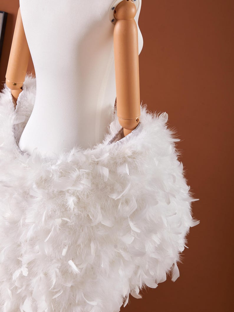 May include: A white, fluffy feather skirt is draped on a mannequin. The skirt is made of soft, white feathers, creating a voluminous and textured appearance. The feathers are densely packed, giving the skirt a full, rounded shape. The background is a solid brown.