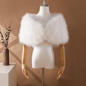May include: A fluffy white feather shawl displayed on a mannequin. The shawl is draped over the shoulders, creating a soft, voluminous look. The mannequin has wooden arms and is set against a neutral background.