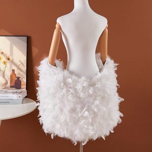 May include: A white feather skirt displayed on a mannequin. The skirt is made of fluffy white feathers and is designed to be worn around the waist. The mannequin has wooden arms and is set against a brown background.