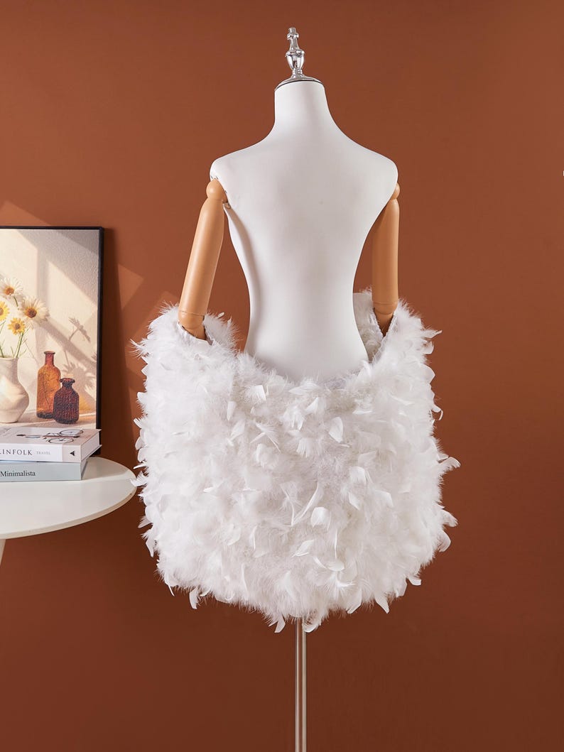 May include: A white feather skirt displayed on a mannequin. The skirt is made of fluffy white feathers and is designed to be worn around the waist. The mannequin has wooden arms and is set against a brown background.