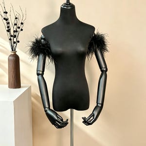 May include: A black off-the-shoulder garment featuring black feather trim along the upper arm sleeves. The piece is displayed on a matte black articulated mannequin torso with posable arms, showcasing a sleek, dark aesthetic with textured plumage.