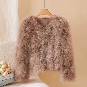 Könnte beinhalten: Staubrosa Jacke mit V-Ausschnitt und langen Ärmeln. Die Jacke besteht aus einem weichen, flauschigen Material, möglicherweise Federn oder Kunstfell, und hängt an einem Holzbügel. Der Hintergrund ist ein neutrales Beige.