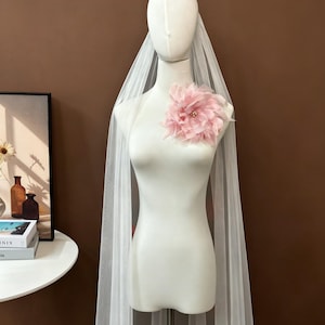 K&ouml;nnte beinhalten: Ein langer, transparenter wei&szlig;er T&uuml;llschleier, der &uuml;ber einer Schaufensterpuppe drapiert ist und im oberen Brustbereich mit einer gro&szlig;en, mehrlagigen, blassrosa Stoffblume verziert ist. Die Blume hat zarte Bl&uuml;tenbl&auml;tter und kleine Perlen.