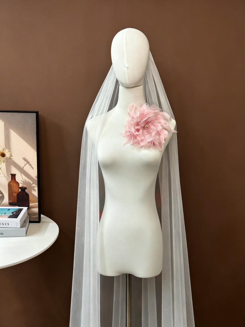 K&ouml;nnte beinhalten: Ein langer, transparenter wei&szlig;er T&uuml;llschleier, der &uuml;ber einer Schaufensterpuppe drapiert ist und im oberen Brustbereich mit einer gro&szlig;en, mehrlagigen, blassrosa Stoffblume verziert ist. Die Blume hat zarte Bl&uuml;tenbl&auml;tter und kleine Perlen.