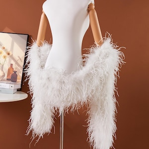 May include: A white feather boa draped over a white mannequin. The boa is made of long, soft, white feathers. The mannequin has wooden arms and is set against a brown background. The boa is a fashion accessory.