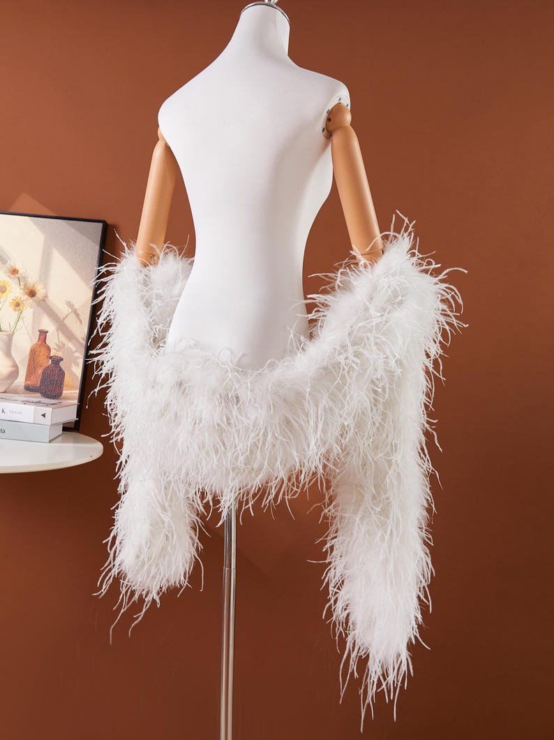 May include: A white feather boa draped over a white mannequin. The boa is made of long, soft, white feathers. The mannequin has wooden arms and is set against a brown background. The boa is a fashion accessory.