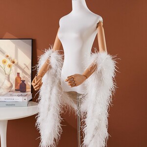May include: A white feather boa draped over a mannequin. The boa is made of long, soft feathers. The mannequin is white with wooden arms and hands. The background is a solid brown color.