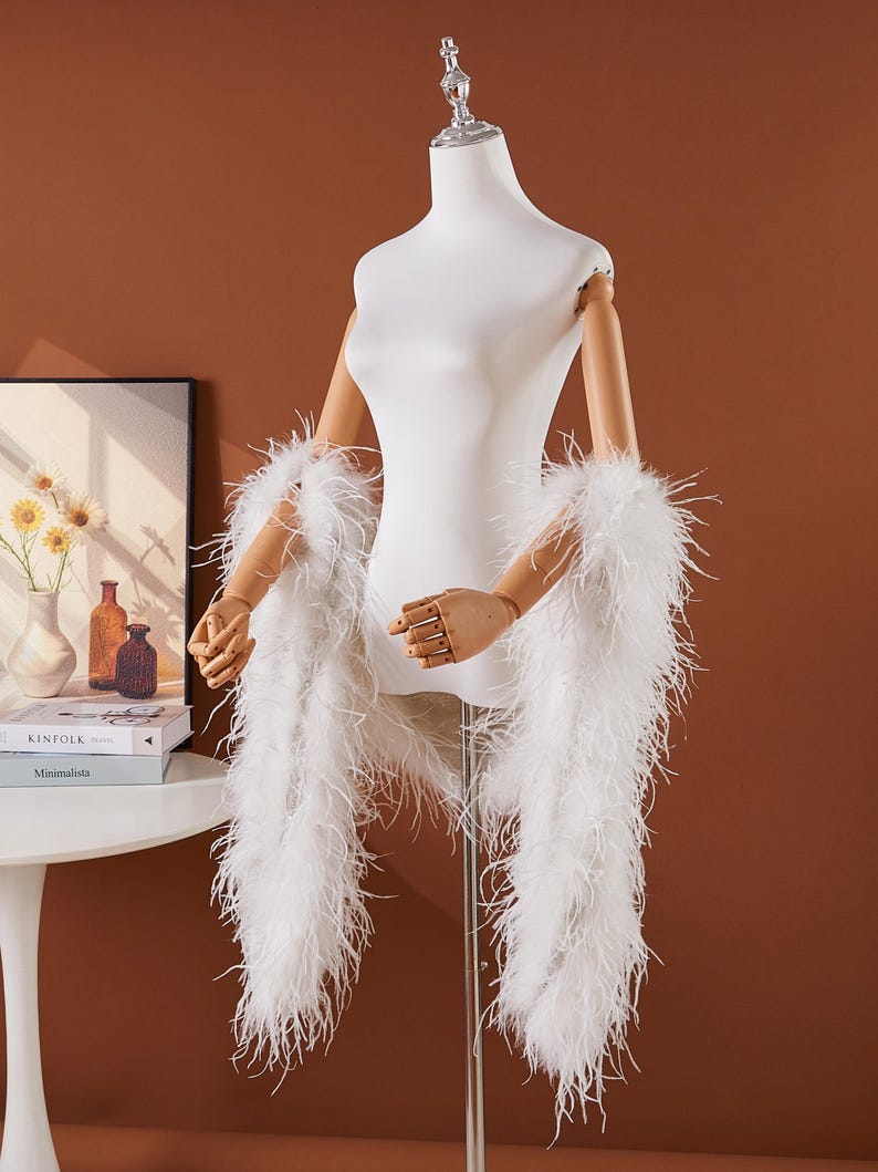 May include: A white feather boa draped over a mannequin. The boa is made of long, soft feathers. The mannequin is white with wooden arms and hands. The background is a solid brown color.