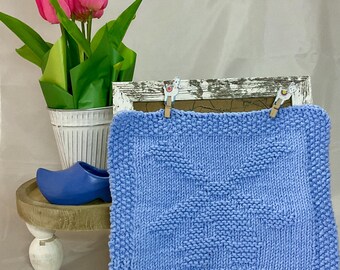 May include: A light blue knitted dishcloth with a geometric pattern is displayed in a white wooden frame. The dishcloth is hanging from two wooden clothespins with llama designs.