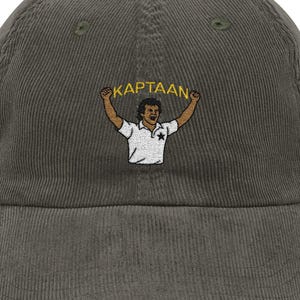 Op de afbeelding: Olijfgroene corduroy baseballcap met een gebogen klep. De cap heeft een geborduurd beeld van een persoon met opgeheven armen, die een banner vasthoudt met de tekst "KAPTAAN". De persoon draagt een wit shirt met een ster.