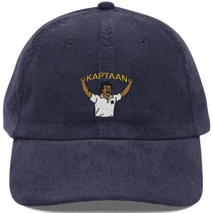 Op de afbeelding: Marineblauwe corduroy baseballcap met een geborduurd beeld van een persoon die een banner vasthoudt met de tekst "KAPTAAN". De persoon draagt een wit shirt. De pet heeft een gebogen klep.