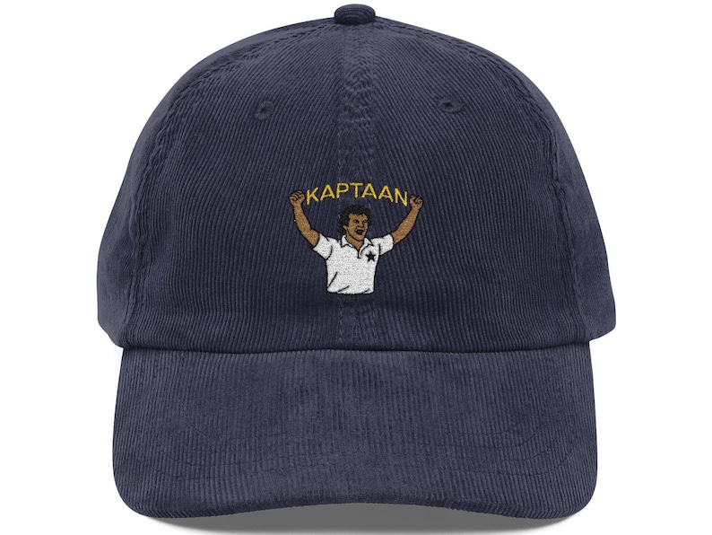 Op de afbeelding: Marineblauwe corduroy baseballcap met een geborduurd beeld van een persoon die een banner vasthoudt met de tekst "KAPTAAN". De persoon draagt een wit shirt. De pet heeft een gebogen klep.