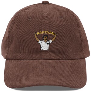 Op de afbeelding: Bruine corduroy baseballcap met een geborduurd ontwerp. Het ontwerp bevat het woord "KAPTAAN" boven een illustratie van een persoon in een wit shirt met opgeheven armen. De cap heeft een gebogen klep en een knoop aan de bovenkant.