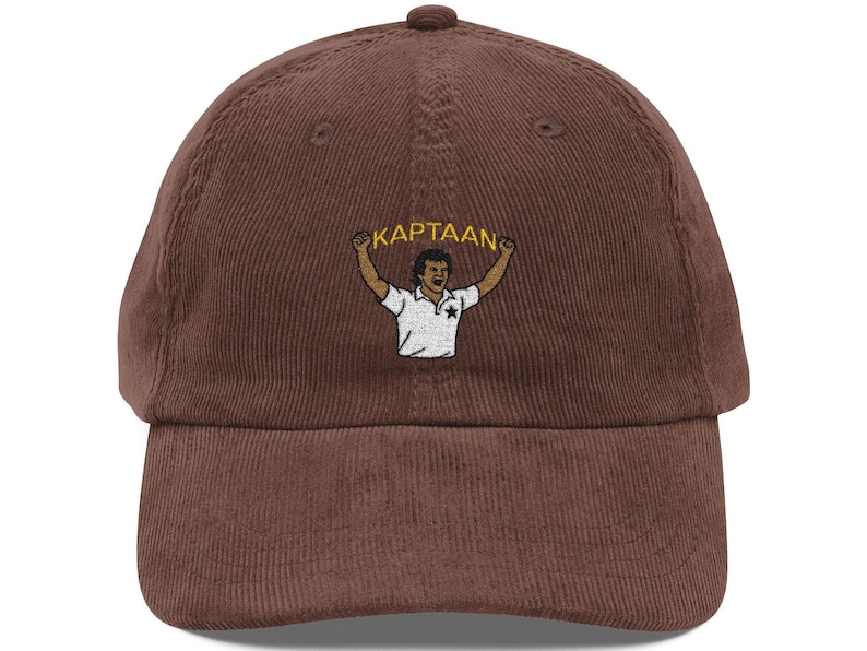 Op de afbeelding: Bruine corduroy baseballcap met een geborduurd ontwerp. Het ontwerp bevat het woord "KAPTAAN" boven een illustratie van een persoon in een wit shirt met opgeheven armen. De cap heeft een gebogen klep en een knoop aan de bovenkant.