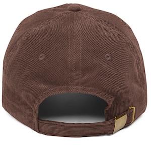 Op de afbeelding: Een bruine corduroy baseballpet met een gebogen klep. De pet heeft een ontwerp met zes panelen, een bovenste knoop en een verstelbare band met een goudkleurige metalen gesp aan de achterkant. De stof heeft een geribbelde textuur.