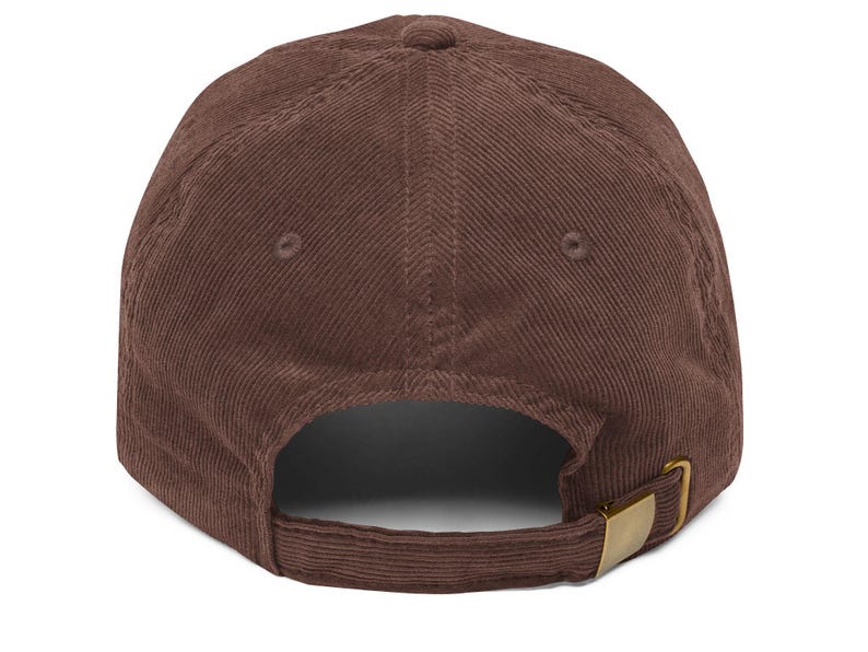 Op de afbeelding: Een bruine corduroy baseballpet met een gebogen klep. De pet heeft een ontwerp met zes panelen, een bovenste knoop en een verstelbare band met een goudkleurige metalen gesp aan de achterkant. De stof heeft een geribbelde textuur.