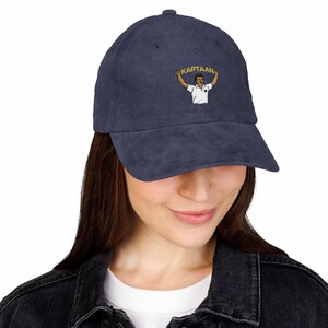 Op de afbeelding: Marineblauwe corduroy baseballcap met een geborduurd ontwerp van een persoon die een banner vasthoudt met het woord "KAPTAAN". De cap heeft een gebogen klep en een gestructureerde kroon. Een modieus accessoire.