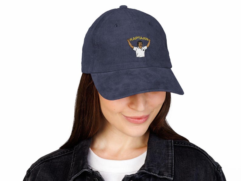 Op de afbeelding: Marineblauwe corduroy baseballcap met een geborduurd ontwerp van een persoon die een banner vasthoudt met het woord "KAPTAAN". De cap heeft een gebogen klep en een gestructureerde kroon. Een modieus accessoire.