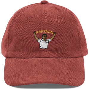 Op de afbeelding: Een baksteenrode corduroy baseballcap met een gebogen klep. De pet heeft een geborduurd ontwerp van een persoon met opgeheven armen en het woord "KAPTAAN" erboven. Het ontwerp is in een contrasterende kleur.