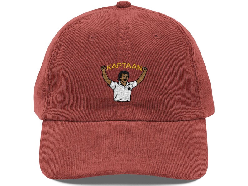 Op de afbeelding: Een baksteenrode corduroy baseballcap met een gebogen klep. De pet heeft een geborduurd ontwerp van een persoon met opgeheven armen en het woord "KAPTAAN" erboven. Het ontwerp is in een contrasterende kleur.
