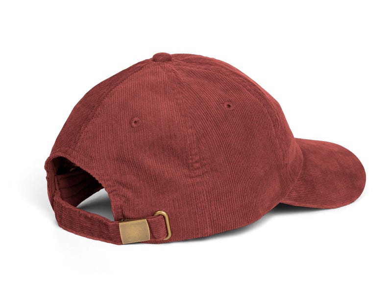 Op de afbeelding: Een bordeauxrode corduroy baseballcap met een gebogen klep. De cap heeft een verstelbare gespsluiting in messingkleur aan de achterkant. De cap is gemaakt van een zachte, getextureerde stof.