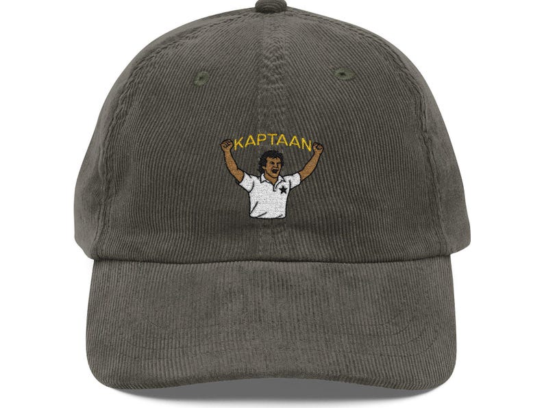 Op de afbeelding: Olijfgroene corduroy baseballcap met een gebogen klep. De cap heeft een geborduurd beeld van een persoon met opgeheven armen, die een banner vasthoudt met de tekst "KAPTAAN". De persoon draagt een wit shirt met een ster.