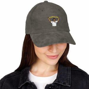 Op de afbeelding: Grijze corduroy baseballcap met een geborduurd beeld van een persoon die een spandoek vasthoudt met het woord "KAPTAAN". De cap heeft een gebogen klep en een stoffen sluiting.