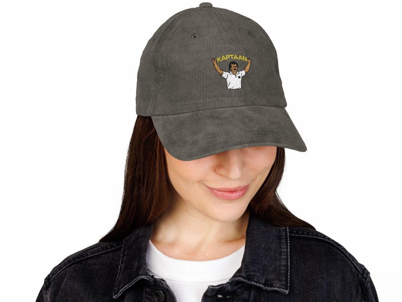 Op de afbeelding: Grijze corduroy baseballcap met een geborduurd beeld van een persoon die een spandoek vasthoudt met het woord "KAPTAAN". De cap heeft een gebogen klep en een stoffen sluiting.