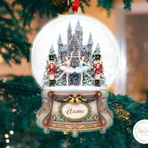 May include: A Christmas ornament snow globe with a ballet theme. The globe features a castle, two ballerinas, and two nutcrackers. The base is decorated with a bow and the word "Name". The ornament hangs from a red ribbon.