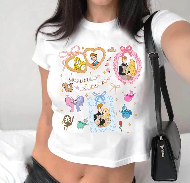 Op de afbeelding: Witte crop top met een grafische print van een cartoonkoppel in een hartvormig frame. De grafiek bevat ook een eekhoorn, een spinnewiel, een strik en de tekst "KUFELU & KATIE".