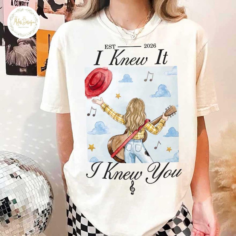 Op de afbeelding: Een cr&egrave;mekleurig t-shirt met een afbeelding van een persoon met lang haar in een geel geruit overhemd en een blauwe spijkerbroek, die een gitaar vasthoudt. Het ontwerp bevat een rode hoed, muzieknoten, sterren, wolken en de tekst "EST 2026 I Knew It I Knew You".