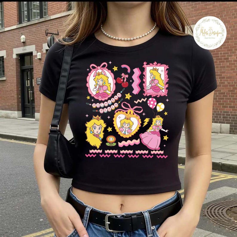 May include: Black crop top featuring multiple cartoon images of Princess Peach in pink frames, hearts, and other shapes. The design includes stars, cherries, and a flower. The top has short sleeves and a round neckline.