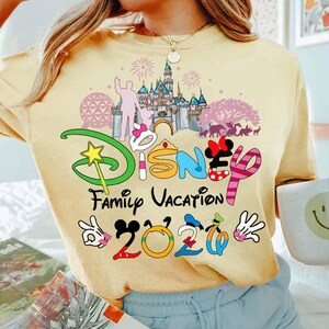May include: A light yellow t-shirt with a colorful Disney Family Vacation 2020 graphic. The graphic includes a castle, fireworks, and Mickey Mouse hands. The shirt is a comfortable, casual style.