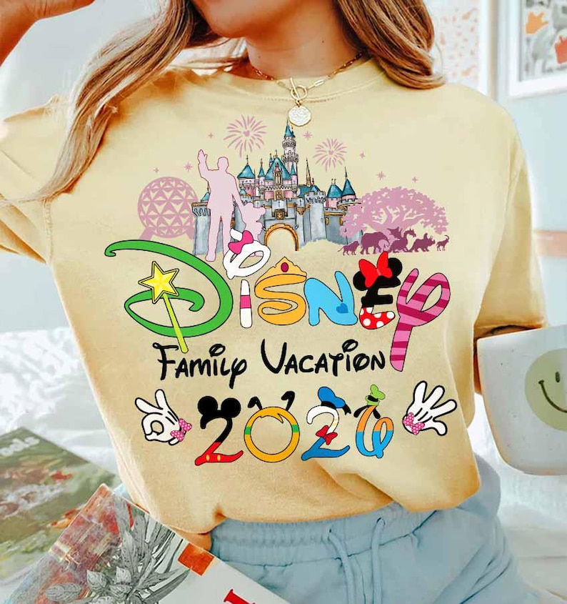 May include: A light yellow t-shirt with a colorful Disney Family Vacation 2020 graphic. The graphic includes a castle, fireworks, and Mickey Mouse hands. The shirt is a comfortable, casual style.