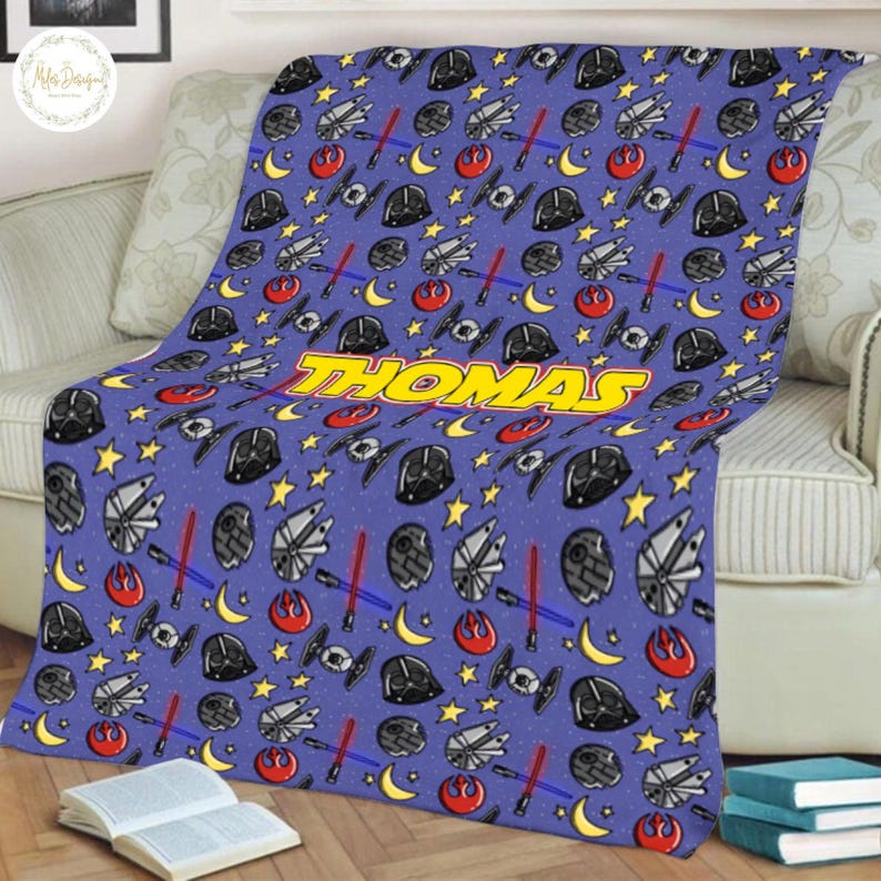 May include: A blue blanket with a repeating pattern of stars, spaceships, and character helmets. The blanket has the name "THOMAS" in yellow block letters. The blanket is draped over a sofa with a book and a stack of books in the foreground.