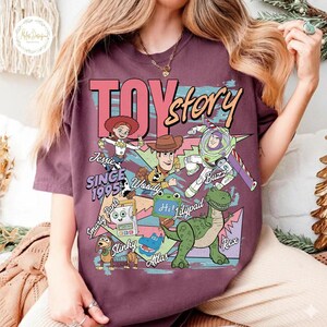 Pode incluir: Uma t-shirt castanha com uma impress&atilde;o gr&aacute;fica das personagens de Toy Story, incluindo Jessie, Woody, Buzz Lightyear, Rex, Slinky, Smarty Pants e Atlas. O texto diz TOY story, SINCE 1995, Jessie, Woody, Buzz, Smarty Pants, Lilypad, Slinky, Atlas e Rex.