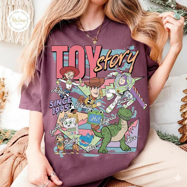 Pode incluir: Uma t-shirt castanha com uma impress&atilde;o gr&aacute;fica das personagens de Toy Story, incluindo Jessie, Woody, Buzz Lightyear, Rex, Slinky, Smarty Pants e Atlas. O texto diz TOY story, SINCE 1995, Jessie, Woody, Buzz, Smarty Pants, Lilypad, Slinky, Atlas e Rex.