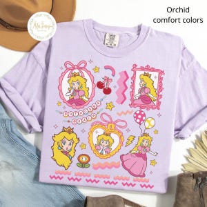 May include: Orchid-colored t-shirt with multiple Princess Peach graphics. The shirt features various illustrations of the character in pink frames, hearts, and other decorative elements. The text "Princess Peach" is also displayed.