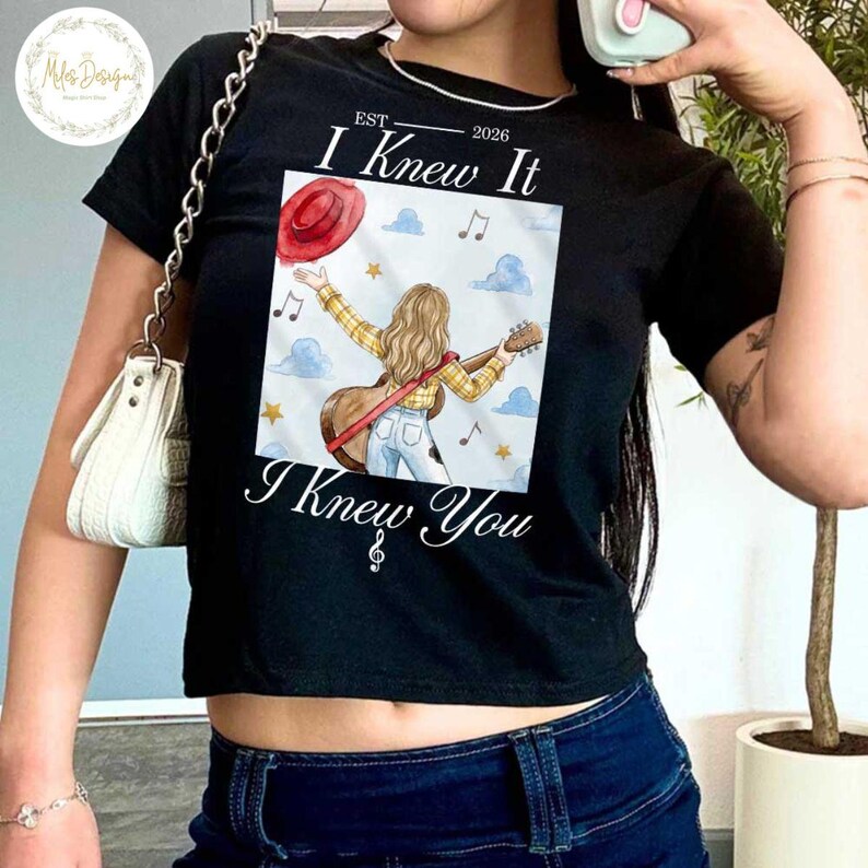 Op de afbeelding: Een zwart T-shirt met korte mouwen en een grafische print van een persoon met lang blond haar in een geel geruit overhemd en een blauwe spijkerbroek, met een gitaar. De tekst luidt "EST 2026 I Knew It I Knew You" met muzieknoten en wolken.