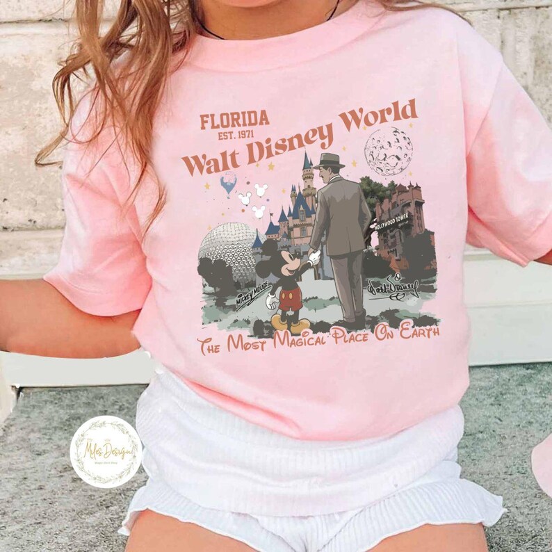 Puede incluir: Camiseta rosa claro con un dise&ntilde;o de Walt Disney World. El dise&ntilde;o incluye el texto "Florida Est. 1971 Walt Disney World" y "The Most Magical Place On Earth". El dise&ntilde;o presenta a Mickey Mouse, un hombre y monumentos del parque.