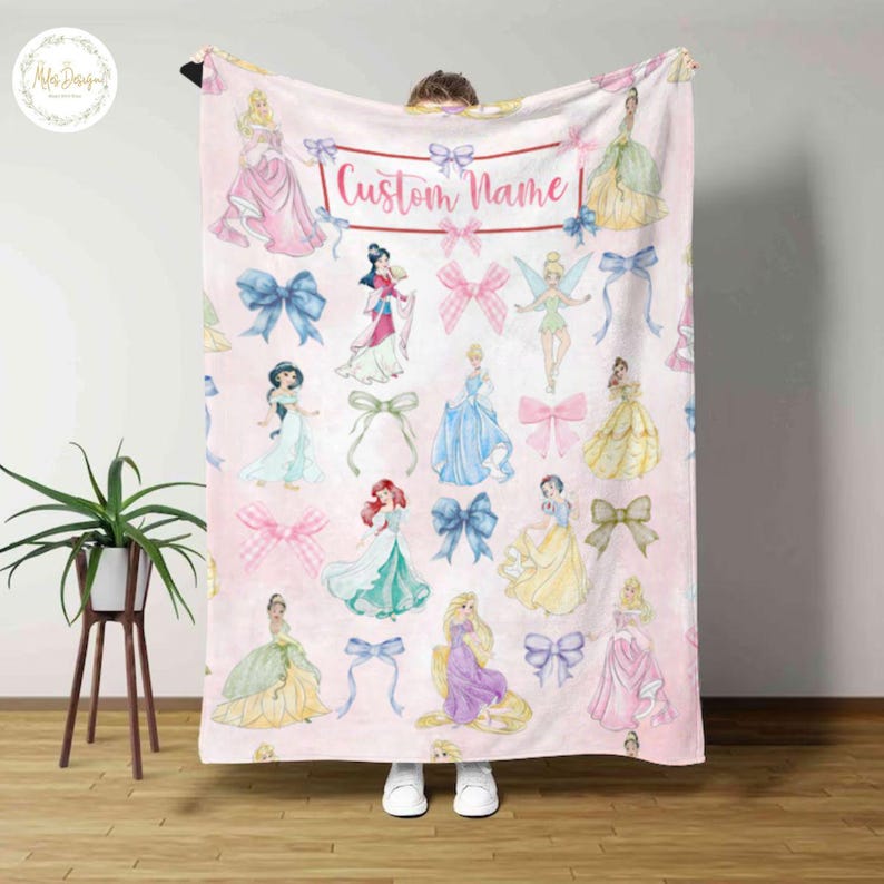 May include: A pink blanket featuring watercolor illustrations of princesses and bows. The blanket has the text "Custom Name" in a rectangular frame. The princesses are in various gowns, and the bows are in shades of blue, pink, and gold.