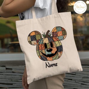 May include: A cream-coloured tote bag with a patchwork pumpkin design and Mickey Mouse ears. The pumpkin is made of various colourful fabric squares and has a smiling face. The word "Name" is written in black script below the pumpkin.