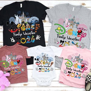 May include: A collection of Disney Family Vacation 2020 t-shirts and a onesie in black, gray, maroon, and pink. The shirts feature Disney characters and a castle graphic. A pink stuffed bunny and checkered slippers are also in the image.