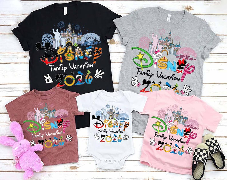 May include: A collection of Disney Family Vacation 2020 t-shirts and a onesie in black, gray, maroon, and pink. The shirts feature Disney characters and a castle graphic. A pink stuffed bunny and checkered slippers are also in the image.