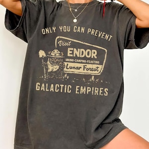 May include: Dark grey t-shirt featuring the text "ONLY YOU CAN PREVENT" above a sign that reads "Visit ENDOR Hiking-Camping-Feasting Lunar Forest GALACTIC EMPIRES". The sign includes a cartoon character with a hat and shovel.
