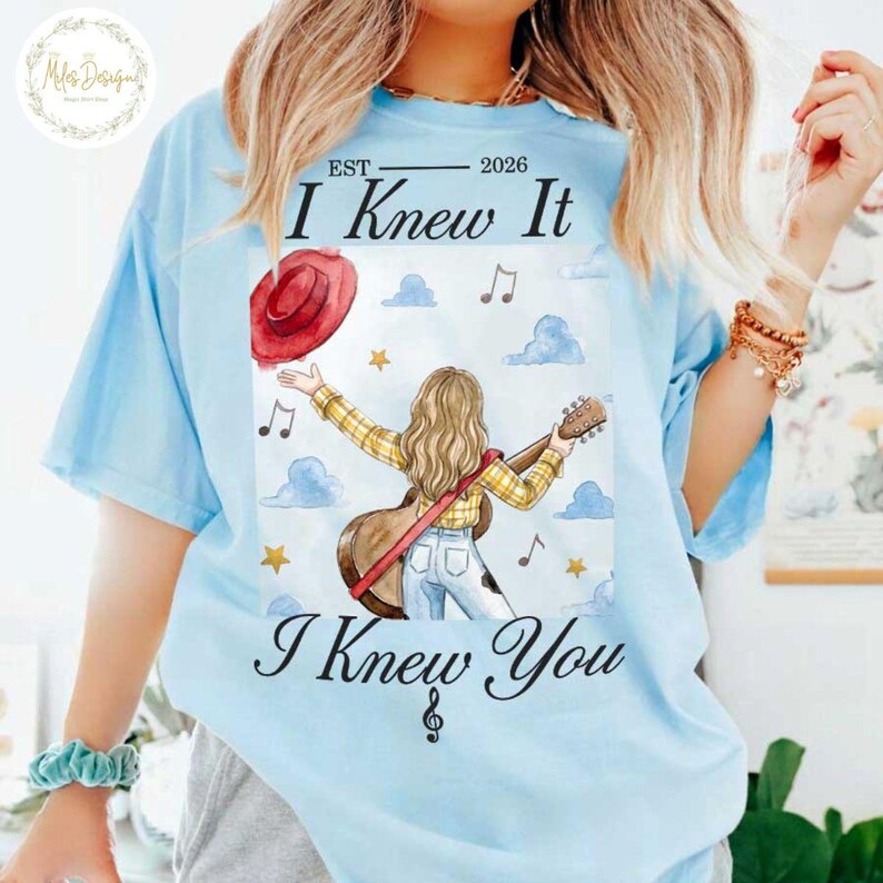 Op de afbeelding: Een lichtblauw t-shirt met een afbeelding van een persoon met lang haar in een geel geruit overhemd en een blauwe spijkerbroek, met een gitaar en een rode hoed. De tekst luidt "EST 2026 I Knew It I Knew You" met muzieknoten, sterren en wolken.
