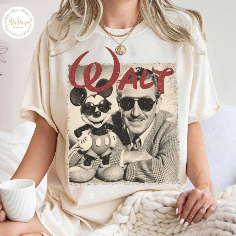 May include: A cream-coloured t-shirt with a vintage graphic of Mickey Mouse and Walt Disney. The graphic includes the word "Walt" in red script. Mickey Mouse is wearing sunglasses. The shirt has short sleeves.