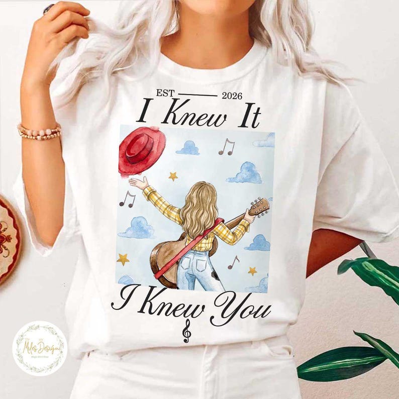 Op de afbeelding: Een wit t-shirt met een grafische illustratie van een persoon met lang blond haar in een geel geruit overhemd en een blauwe spijkerbroek, met een gitaar. De tekst luidt EST 2026 I Knew It I Knew You, met een rode hoed en muzieknoten.